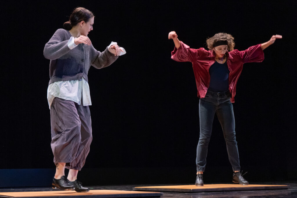 Two dancers, one in gray and one in red and black, in tap shoes on square tap boards. The dancer in red (me) has arms extended and is slightly airborne.