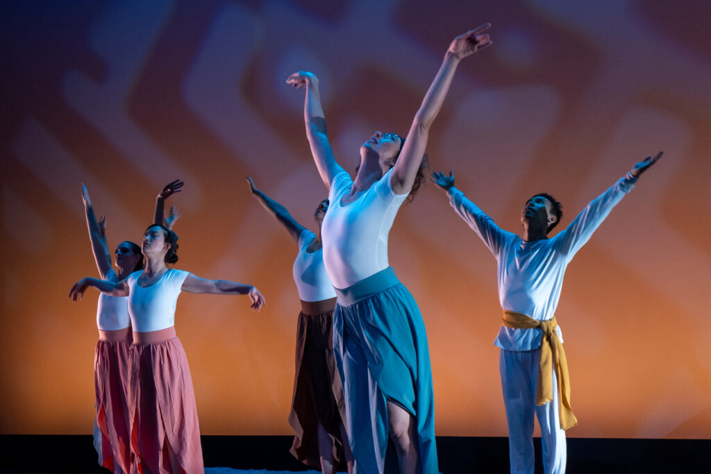 Five dancers with arms extended and heads tilted to the sky. The closest dancer (me) is in a white top and blue flowing pants. Three dancers behind wear white tops and pink flowing pants. One wears all white with a yellow sash.