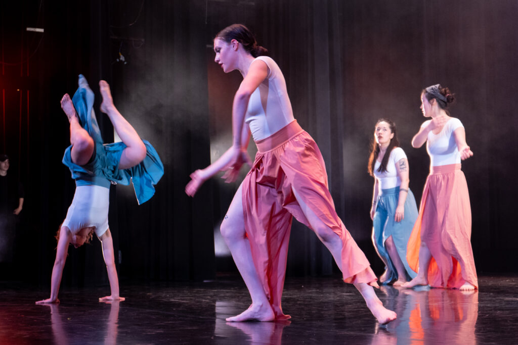 Four dancers on stage. Two are dressed in white tops with blue flowing pants and two are in white tops with pink flowing pants. The closest dancer in pink leans and reaches one arm. The closest dancer in blue (me) is in a handstand with legs folded.