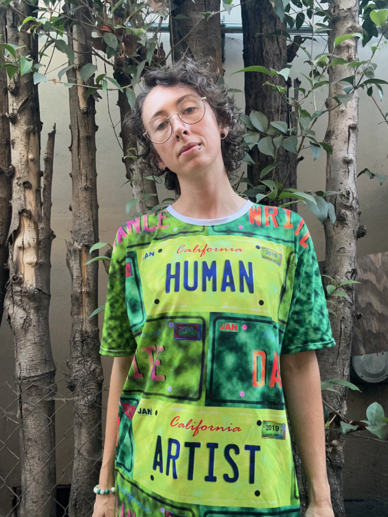 A person with brown curly hair and blue eyes wearing silver glasses, a black lip ring, and a t-shirt top in shades of green. The images on the tank top resemble license plates, and read HUMAN ARTIST. Behind the individual are several leafy trees and a concrete wall.