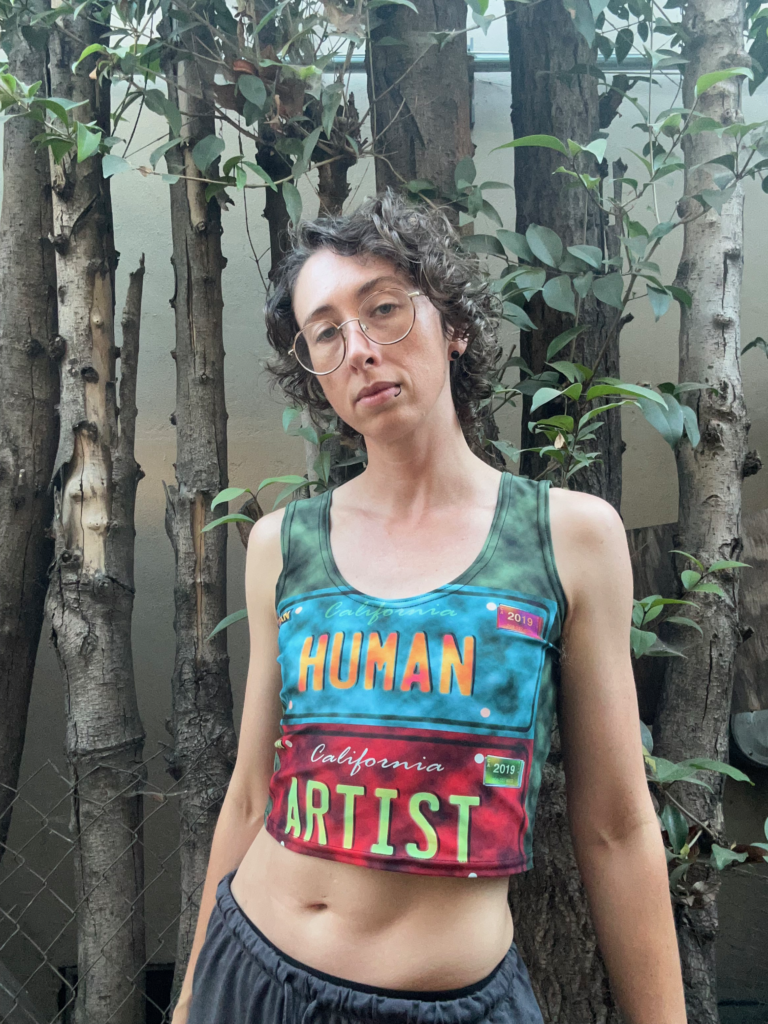 A person with brown curly hair and blue eyes wearing silver glasses, a black lip ring, and a cropped tank top in green, blue, and red. The images on the tank top resemble license plates, with a blue one reading HUMAN in orange text and a red one reading ARTIST in green text. Behind this individual are several leafy trees and a concrete wall.